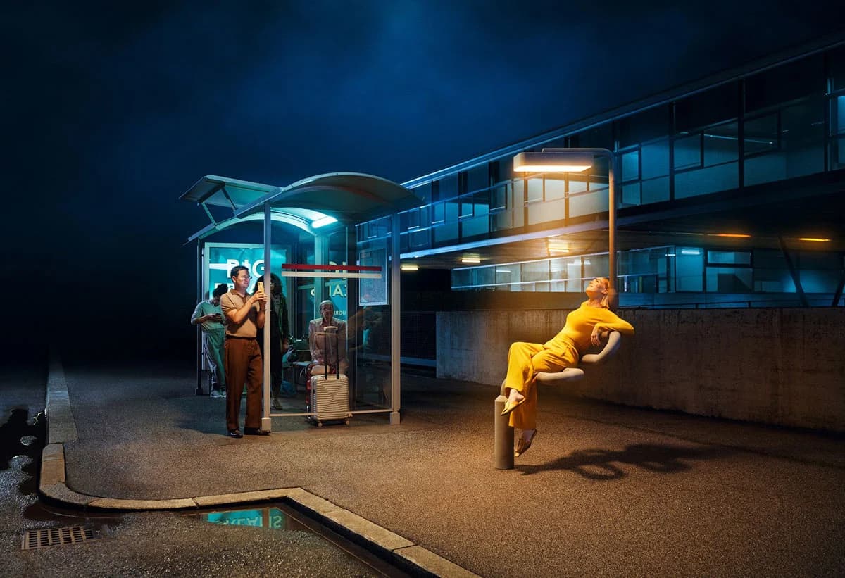 Eine Person in gelber Kleidung sitzt nachts auf einem Pfosten in der Nähe einer Bushaltestelle, andere warten unter einem Unterstand.