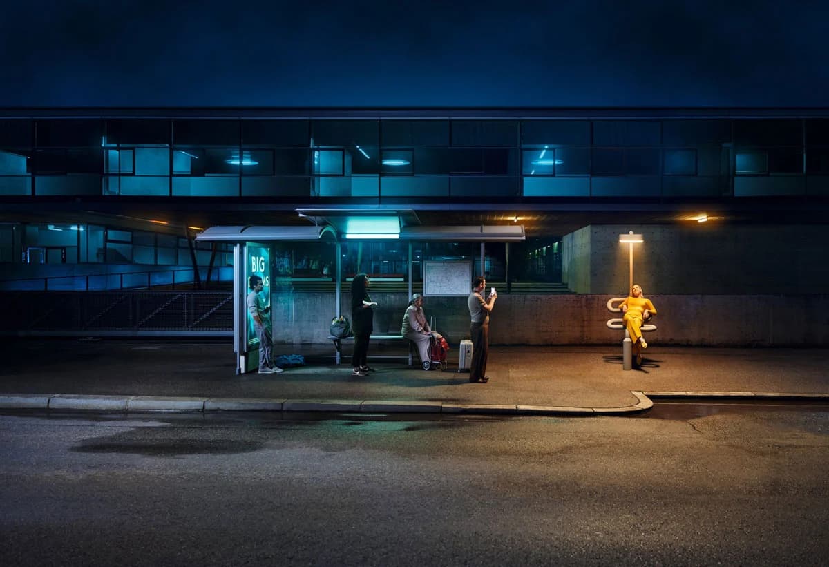 Menschen warten nachts an einer Bushaltestelle, eine Person im gelben Outfit sitzt abseits unter einer Laterne.