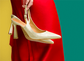 Escarpins à talons beiges tenus à la main devant une robe rouge sur fond jaune et vert