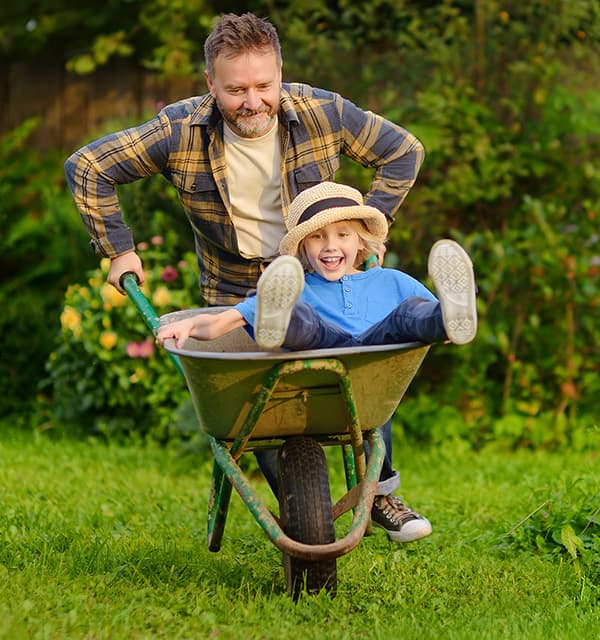 Erwachsener schiebt Kind im Sonnenhut in einer Schubkarre über eine grüne Wiese im Garten