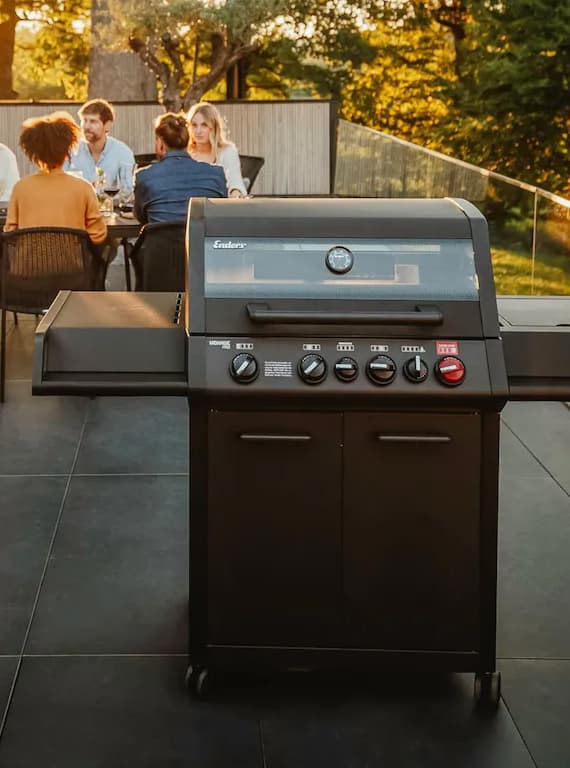 Großer Gasgrill auf Terrasse, im Hintergrund sitzt eine Gruppe Menschen gemeinsam am Esstisch draußen