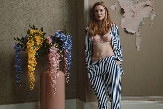 Femme debout en soutien-gorge à fleurs et pantalon rayé bleu et blanc, près d’un grand vase de fleurs colorées