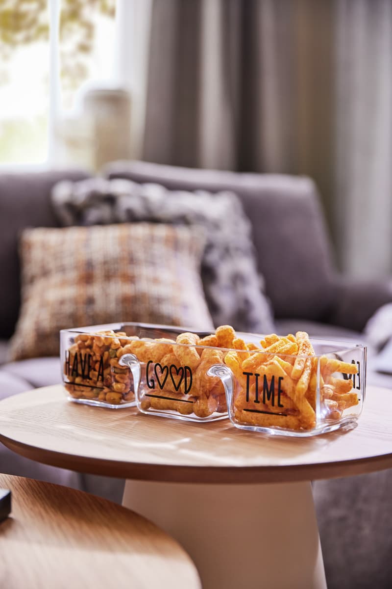 Glasbehälter mit Text "Have a good time" gefüllt mit Snacks auf Holztisch, Couch und Kissen im Hintergrund