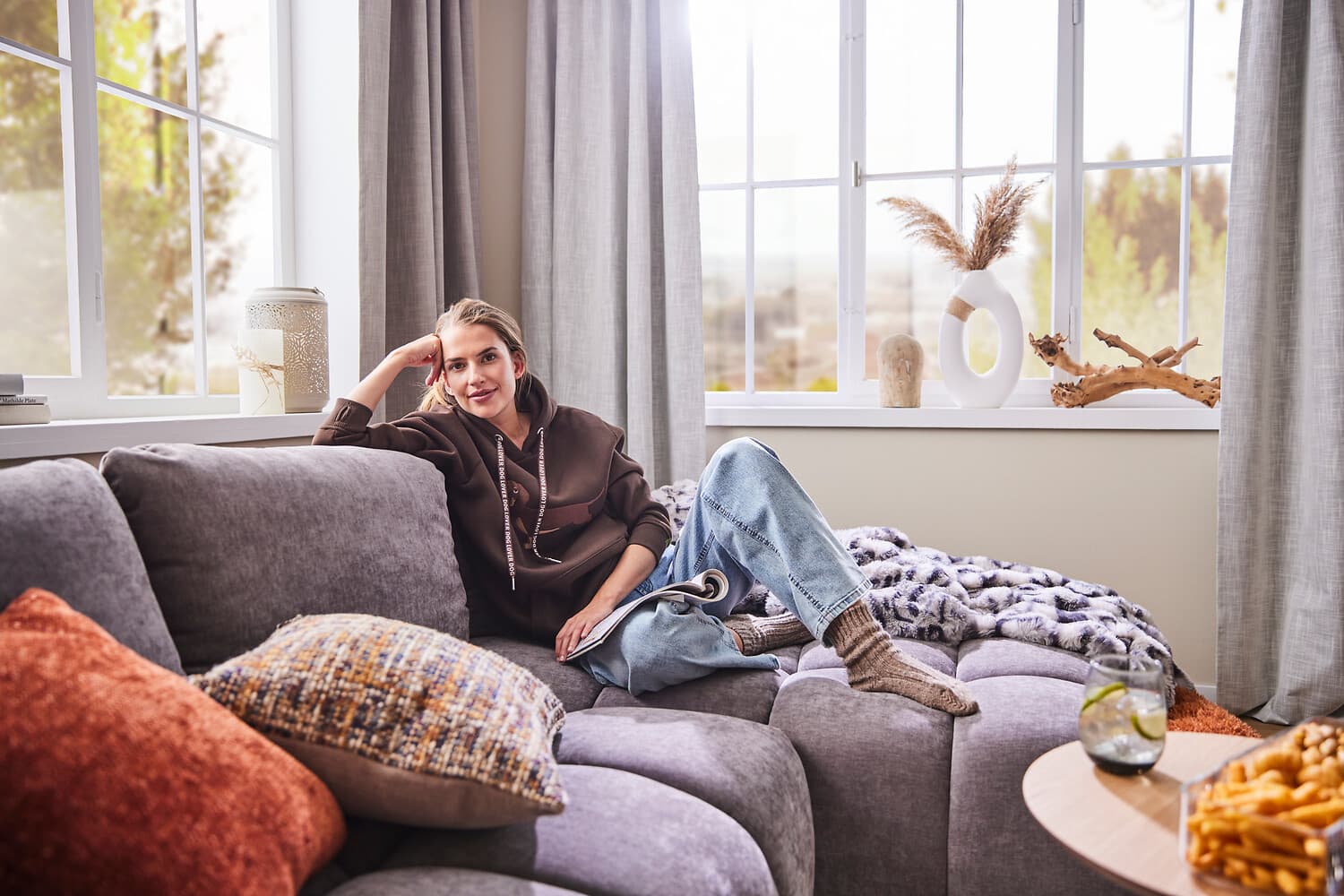 Gemütliches Wohnzimmer mit grauem Sofa, bunten Kissen und zwei runden Beistelltischen vor einem großen Fenster mit Vorhängen