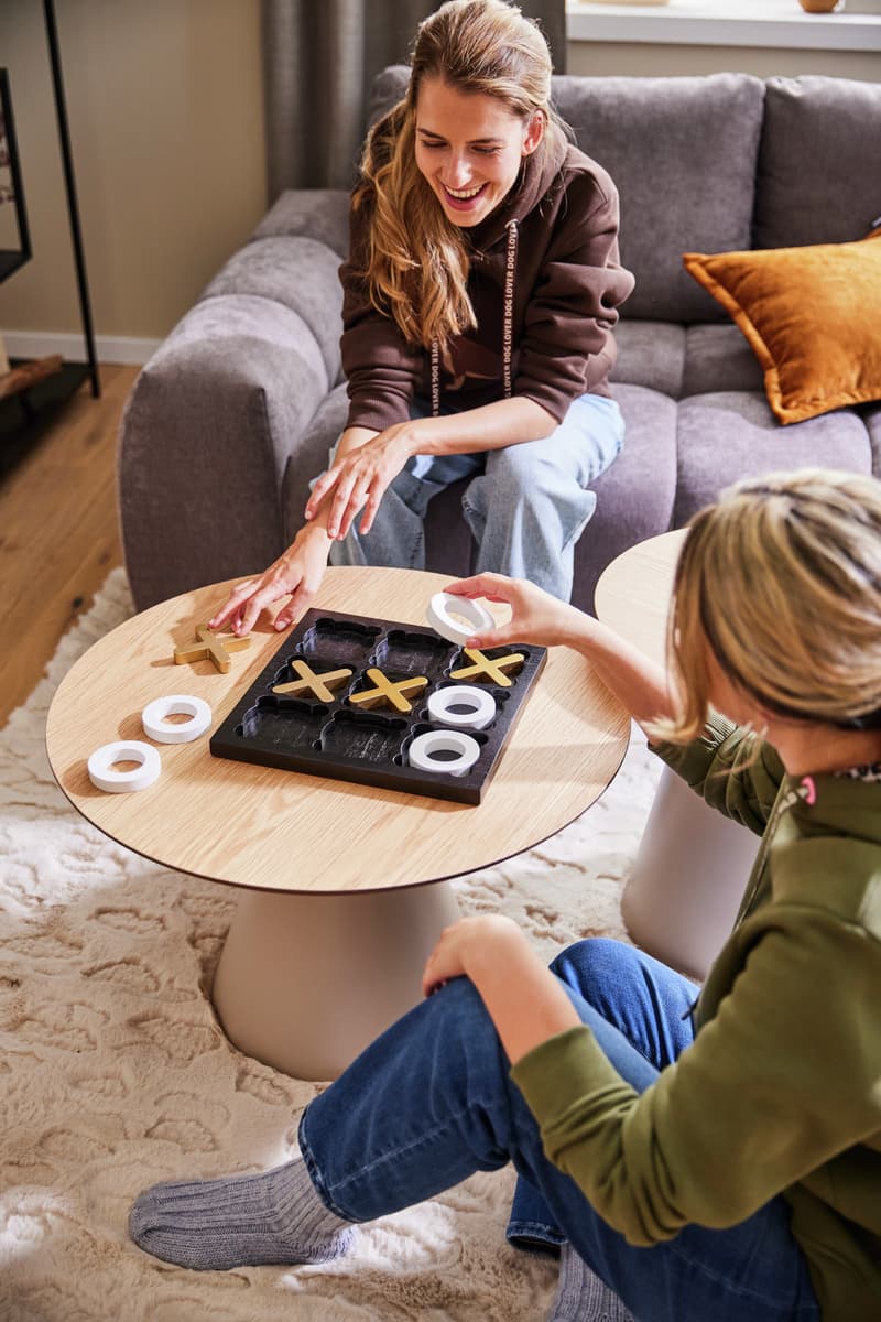Zwei runde Beistelltische mit Spiel und Snacks darauf, im Hintergrund graues Sofa mit bunten Kissen und Fenster mit Vorhängen.