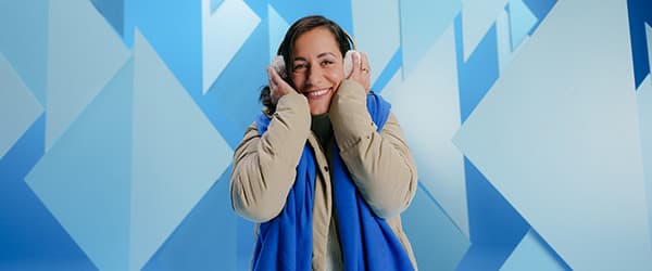 Person in beiger Jacke und blauem Schal hält weiße Kopfhörer vor einem abstrakten blauen Hintergrund