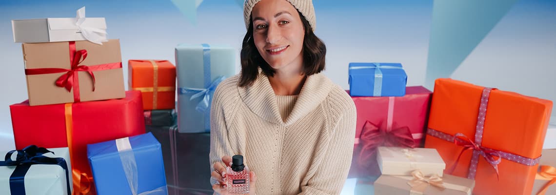 Frau in beige Strickpullover hält Parfümflasche, sitzt vor bunten Geschenkboxen mit Schleifen.