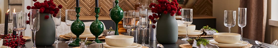 Eleganter Esstisch mit roten Blumen, grünen Kerzenhaltern, weißen Tellern, Gläsern und Besteck für festliches Dinner gedeckt.