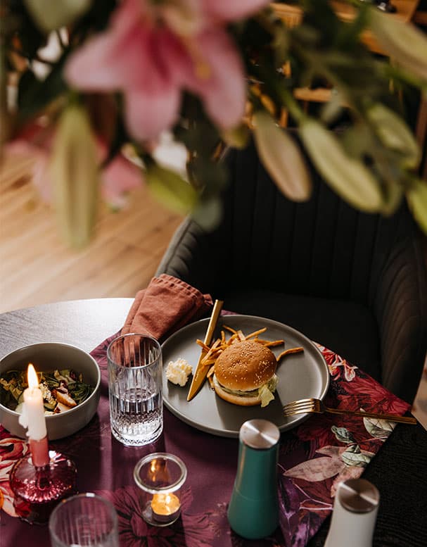 Beleuchteter Tisch mit Burger, Pommes, Salat und Getränk auf floralem Tuch. Kerze und Blume im Vordergrund.
