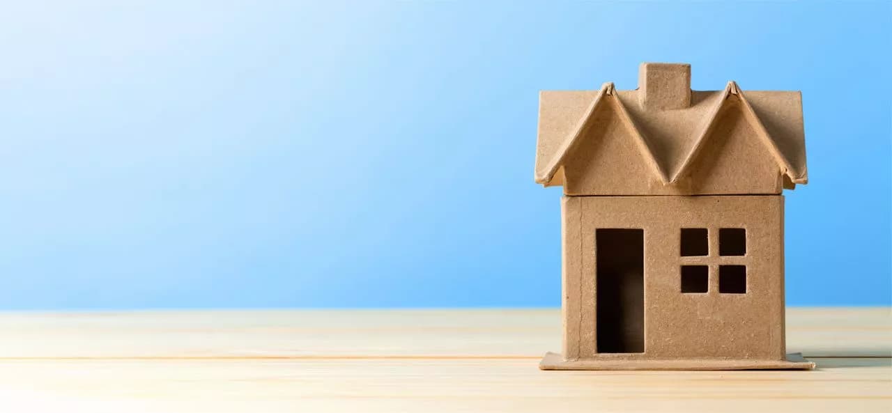 Papierhausmodell mit offenem Fenster und Tür auf einem Holztisch vor blauem Hintergrund