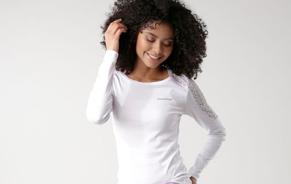 Frau in weißem Langarmshirt berührt ihr lockiges Haar und steht seitlich.
