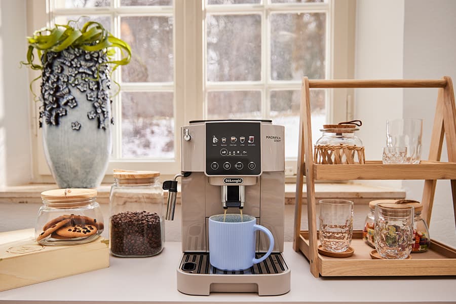 Kaffeevollautomat bereitet Kaffee in blauer Tasse zu, daneben Kekse, Kaffeebohnen und Gläser in Regalen.