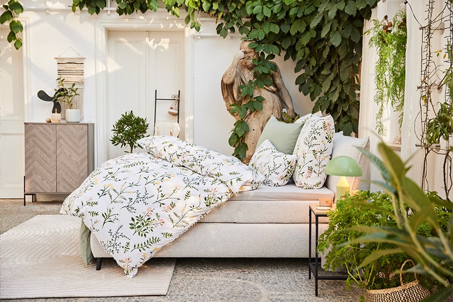 Gemütliches Schlafzimmer mit Bettwäsche im floralen Design, umgeben von Pflanzen und Dekorationen in einem hellen Raum.