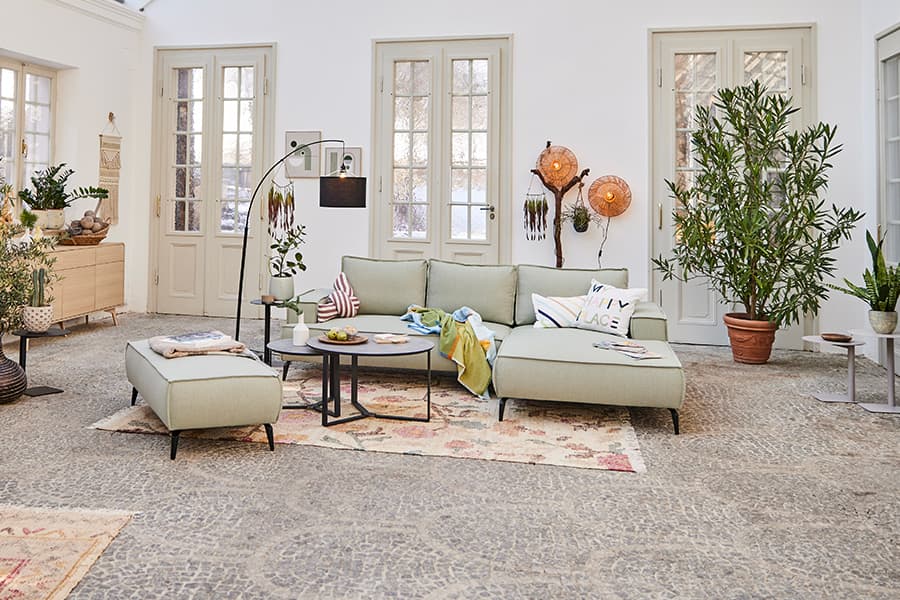 Helles Wohnzimmer mit grüner Eckcouch, Couchtischen und Pflanzen vor großen Fenstern.