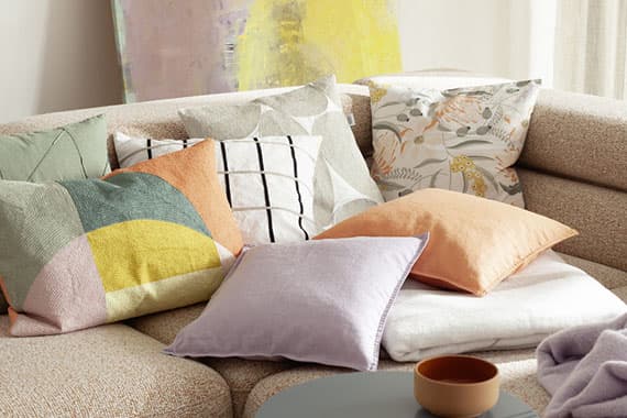 Verschiedene bunte Kissen auf einem beigefarbenen Sofa, im Hintergrund ein abstraktes Gemälde.