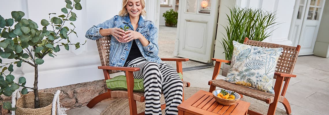 Frau im gestreiften Lounge-Outfit sitzt mit Getränk auf Stuhl neben kleinem Holztisch und zweitem Stuhl mit Zierkissen