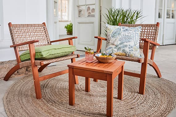 Zwei Holzstühle mit Kissen und kleinem Tisch auf rundem Teppich in hellem Raum mit Pflanzen.