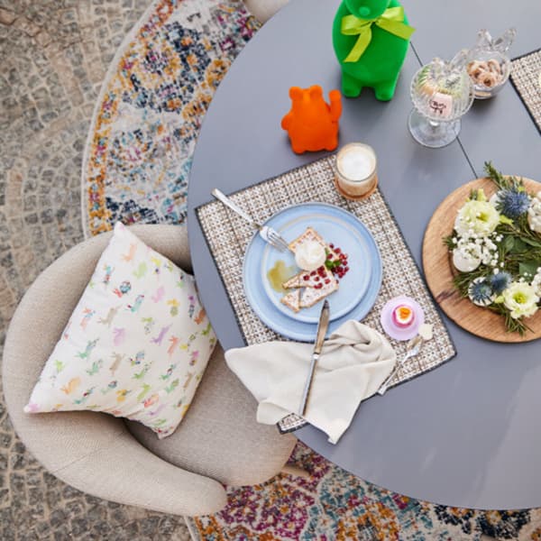 Gedeckter Tisch mit Teller, Besteck und Dessert. Dekoration mit Blumen und bunten Figuren. Kissen auf Stuhl.