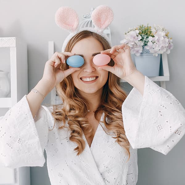 Personne en peignoir blanc avec serre-tête oreilles de lapin roses, posant devant une étagère décorée de fleurs.