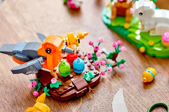 Bunte LEGO-Vögel und Blumen auf einem Holztisch, im Hintergrund LEGO-Hase und Szenerie.