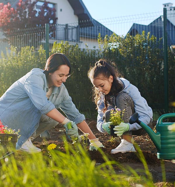 Zwei Personen knien im Gartenbeet und pflanzen Blumen, neben ihnen steht eine grüne Gießkanne