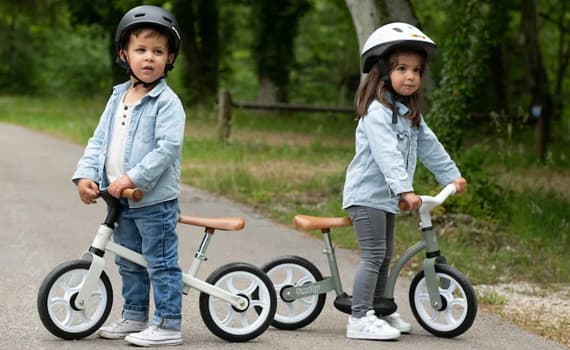 Zwei Kinder mit Helmen stehen im Park auf Laufrädern und halten den Lenker auf einem Weg im Grünen