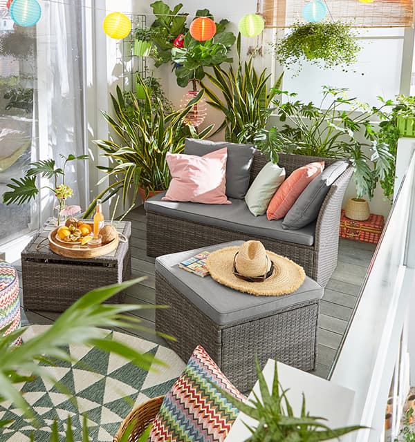 Gemütliche Balkonlounge mit grauem Sofa, bunten Kissen, Pflanzen, Lichterkette, Tablett mit Getränken und Strohhut auf Hocker