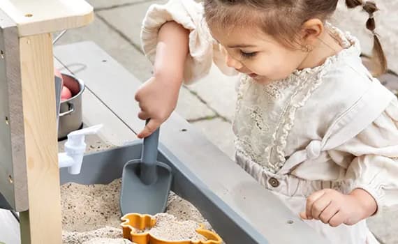Kind spielt an Sandtisch, schaufelt Sand in Form, daneben kleine Spielküche mit Töpfen