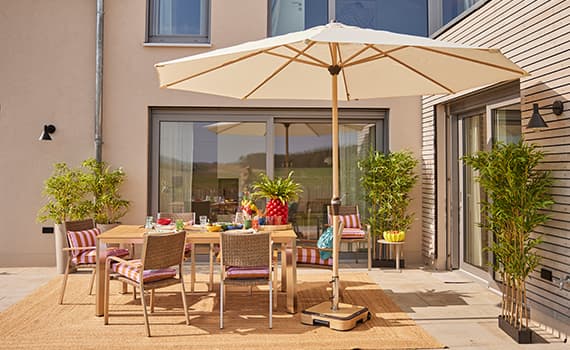 Paravent auf sonniger Terrasse, umgeben von Pflanzen, davor Sitzgruppe mit Tisch, Getränken und Lampe