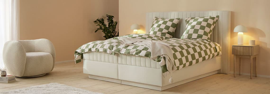 Doppelbett mit grün kariertem Bettbezug, zwei Kissen, Bank am Fußende, Nachttische mit Lampen in hellem Schlafzimmer