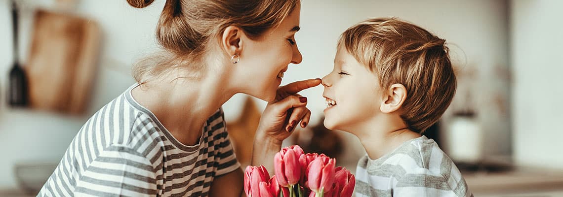 Frau und Kind in gestreiften Shirts berühren sich lächelnd die Nasen, vorn steht ein Strauß pinker Tulpen