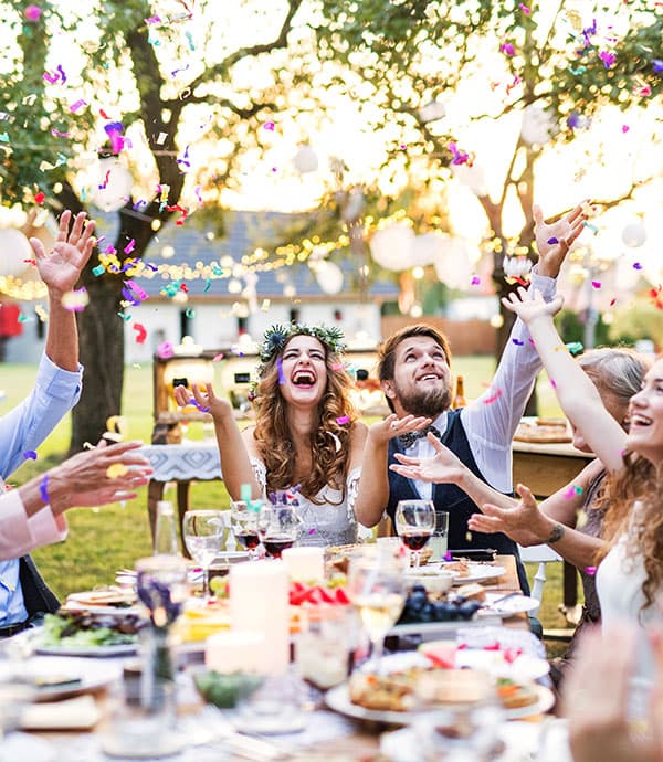Hochzeitsgesellschaft sitzt an gedeckter Tafel im Garten und wirft bunte Konfetti in die Luft