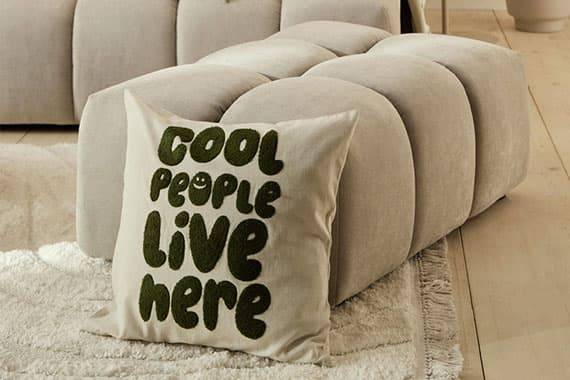 Kissen mit Schriftzug „Cool people live here“ steht auf Teppich vor gepolstertem Hocker im Wohnzimmer