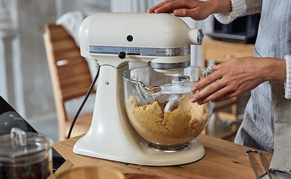 Person bedient Küchenmaschine auf Arbeitsplatte, im Glasrührbehälter wird ein Teig gemischt