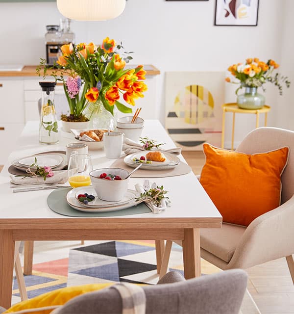 Frühstückstisch mit Müsli, Croissants und Saft, umgeben von bunten Blumensträußen und Stühlen mit Kissen