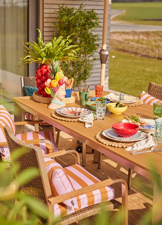Sommerlich gedeckter Gartentisch mit buntem Geschirr, gestreiften Polsterstühlen und großer Obstdeko als Mittelpunkt