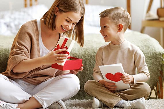 Frau sitzt mit Kind auf dem Boden, öffnet Geschenkbox, Kind hält Karte mit rotem Herz in den Händen