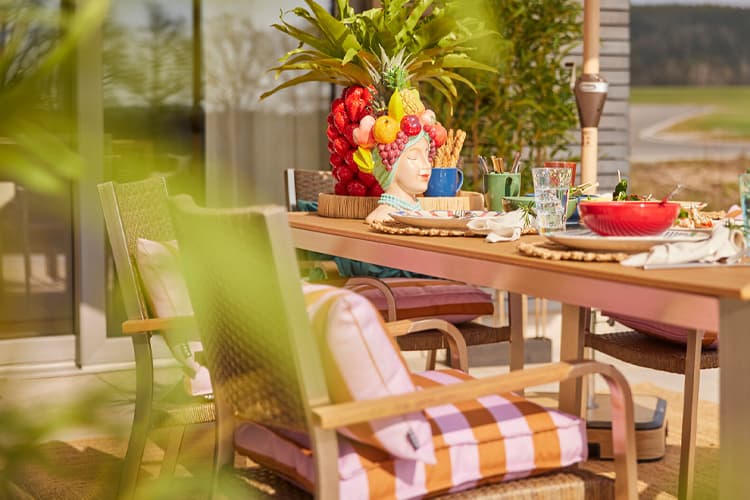 Table de jardin dressée avec vaisselle colorée, verres, coussins rayés et grande composition de fruits au centre