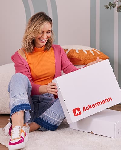Ackermann Paket – sitzende Person öffnet weißen Versandkarton im Wohnzimmer mit Deko und Blumenständer