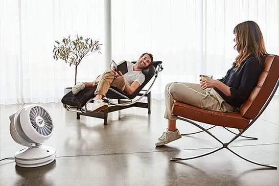 Ventilator auf dem Boden kühlt zwei Personen, die entspannt in Sesseln im hellen Wohnzimmer sitzen