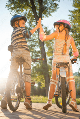 Zwei Kinder mit Helmen geben sich auf Fahrrädern ein High Five auf einem sonnigen Weg im Park