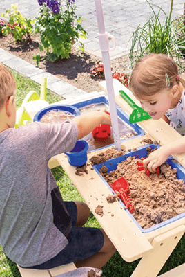 Zwei Kinder spielen im Garten an einem Sand- und Wassertisch mit Eimern, Förmchen und Schaufeln
