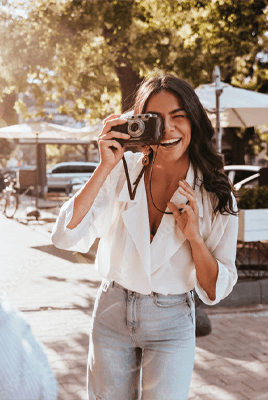 Frau im hellen Oberteil und Jeans fotografiert im Sonnenschein eine Person auf einer belebten Straße