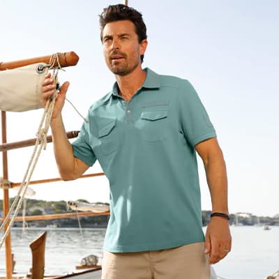 Mann im türkisfarbenen Poloshirt mit Brusttaschen steht auf einem Boot und hält ein Seil, Wasser und Küste im Hintergrund