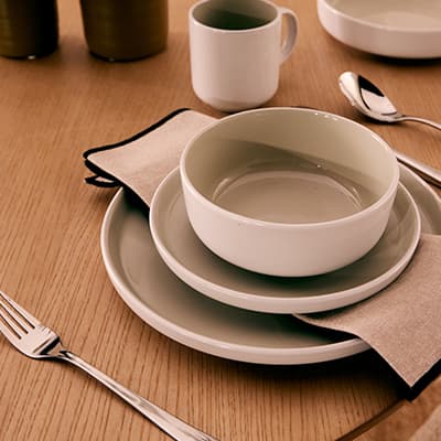 Tischgedeck mit Schüssel auf zwei Tellern, Serviette dazwischen, daneben Besteck und Tasse auf Holzoberfläche