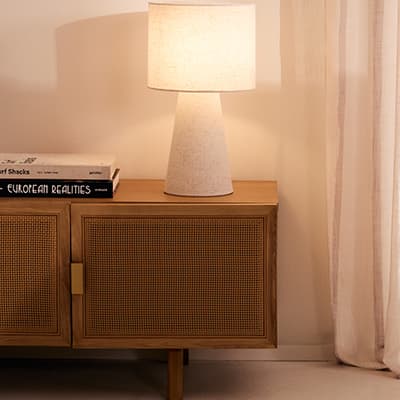 Tischlampe auf Sideboard neben Büchern, warmes Licht vor hellem Vorhang in gemütlicher Zimmerecke