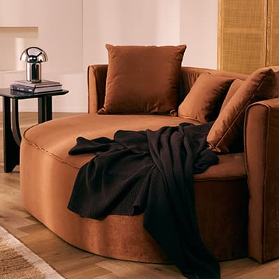 Rundes braunes Sofa mit zwei Kissen, dunkle Decke lässig über Sitzfläche, daneben Beistelltisch mit Lampe