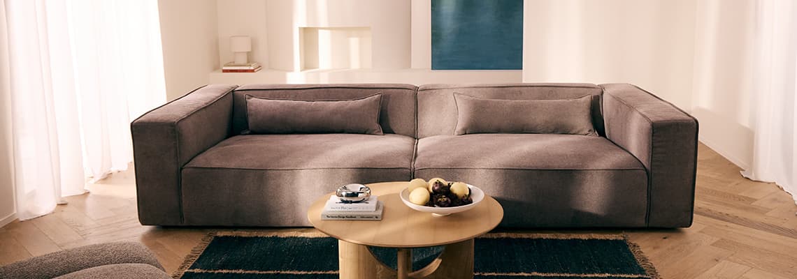 Großes graues Sofa vor heller Wand, runder Couchtisch mit Obstschale und Büchern, dahinter Kamin und blaues Wandbild