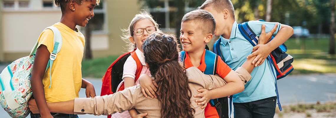 Frau vor Schule umarmt zwei lachende Mädchen mit Rucksack und Heften auf dem Schulhof