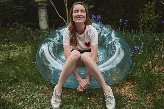 Frau im T-Shirt mit rotem Herz sitzt lächelnd auf durchsichtigem Sitz im Garten mit Blumen im Hintergrund